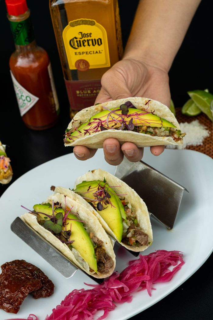 Hand holding tacos with avocado, Tabasco, and tequila on a plate.