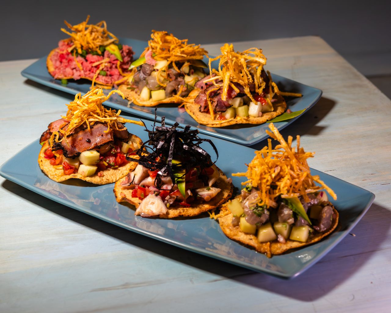 A selection of gourmet tostadas topped with meat, vegetables, and crispy garnishes, served on blue plates.