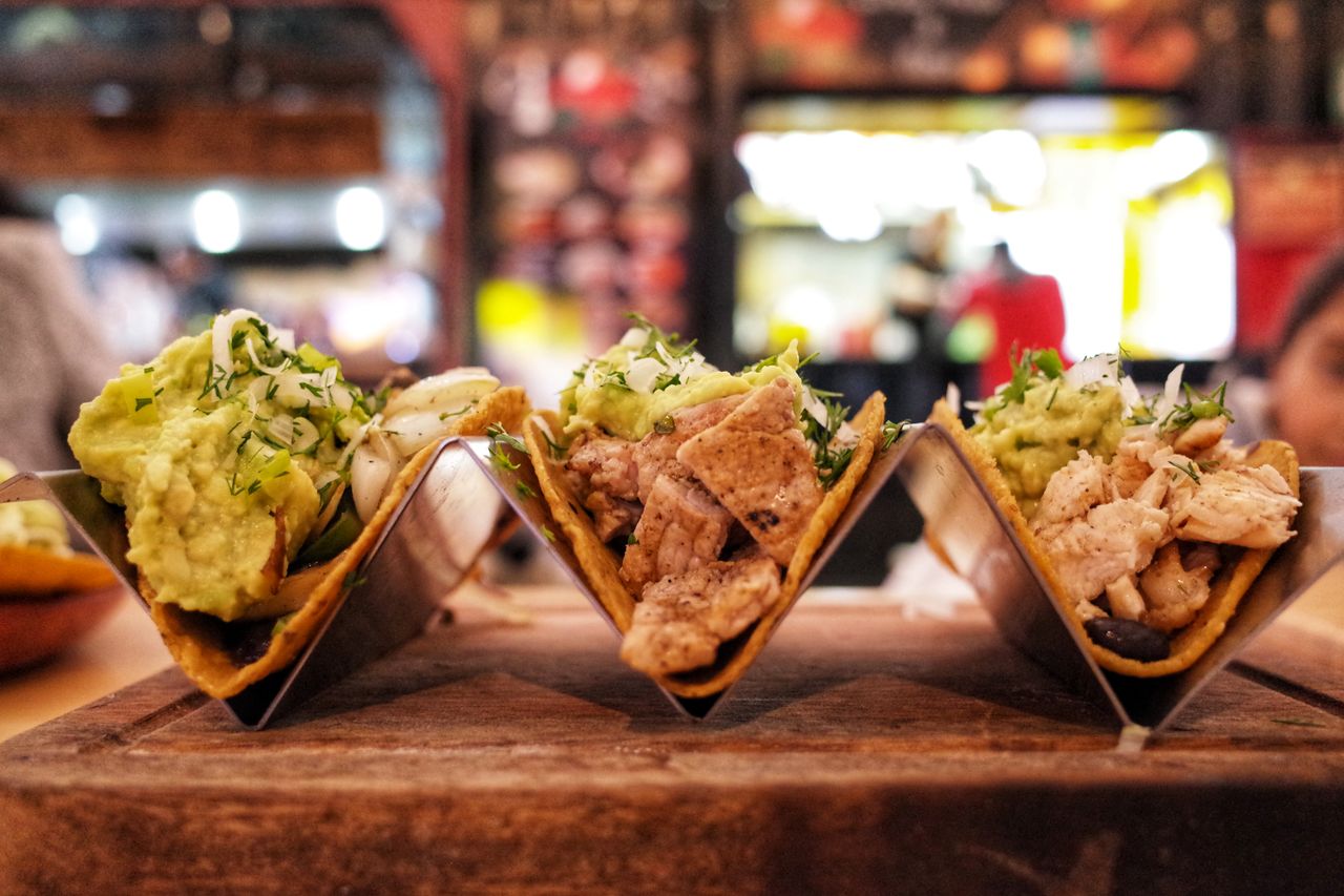 gallery-05 Close-up of three chicken tacos with guacamole, herbs, and onions in a restaurant setting.